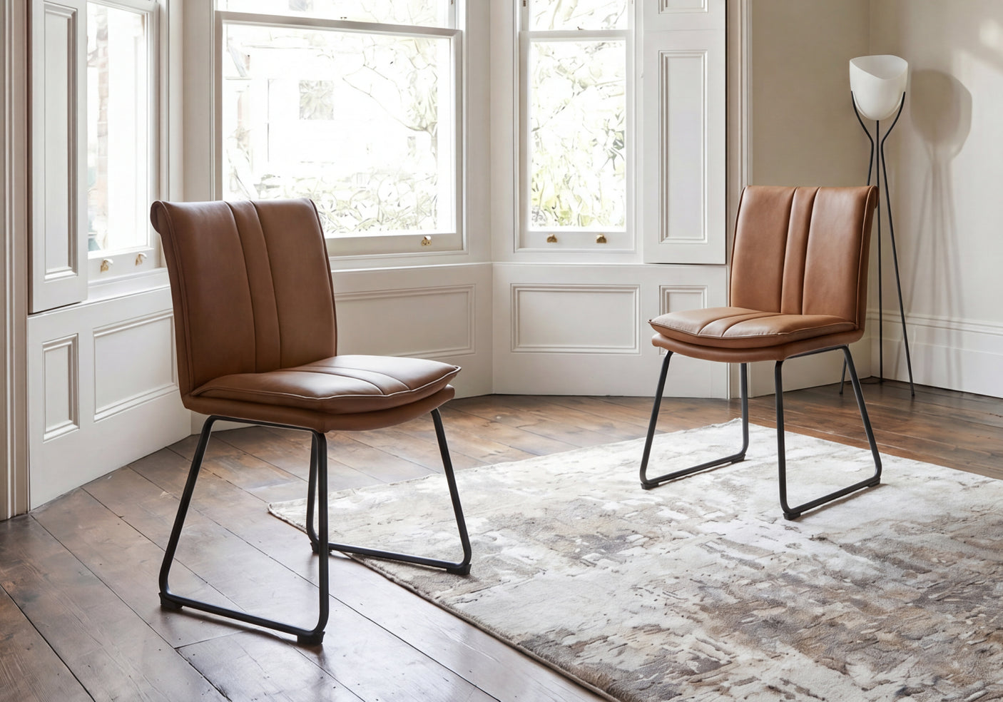 Two brown leather chairs in a room with large victorian windows and a rug.
